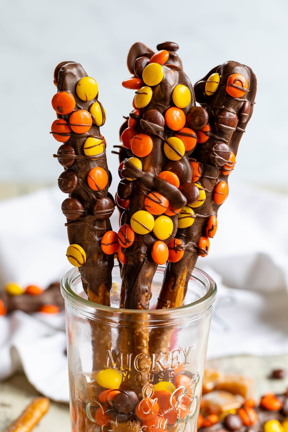 Three pretzel rods dipped in chocolate and coated with orange, yellow, and brown candy pieces are standing upright in a glass jar.