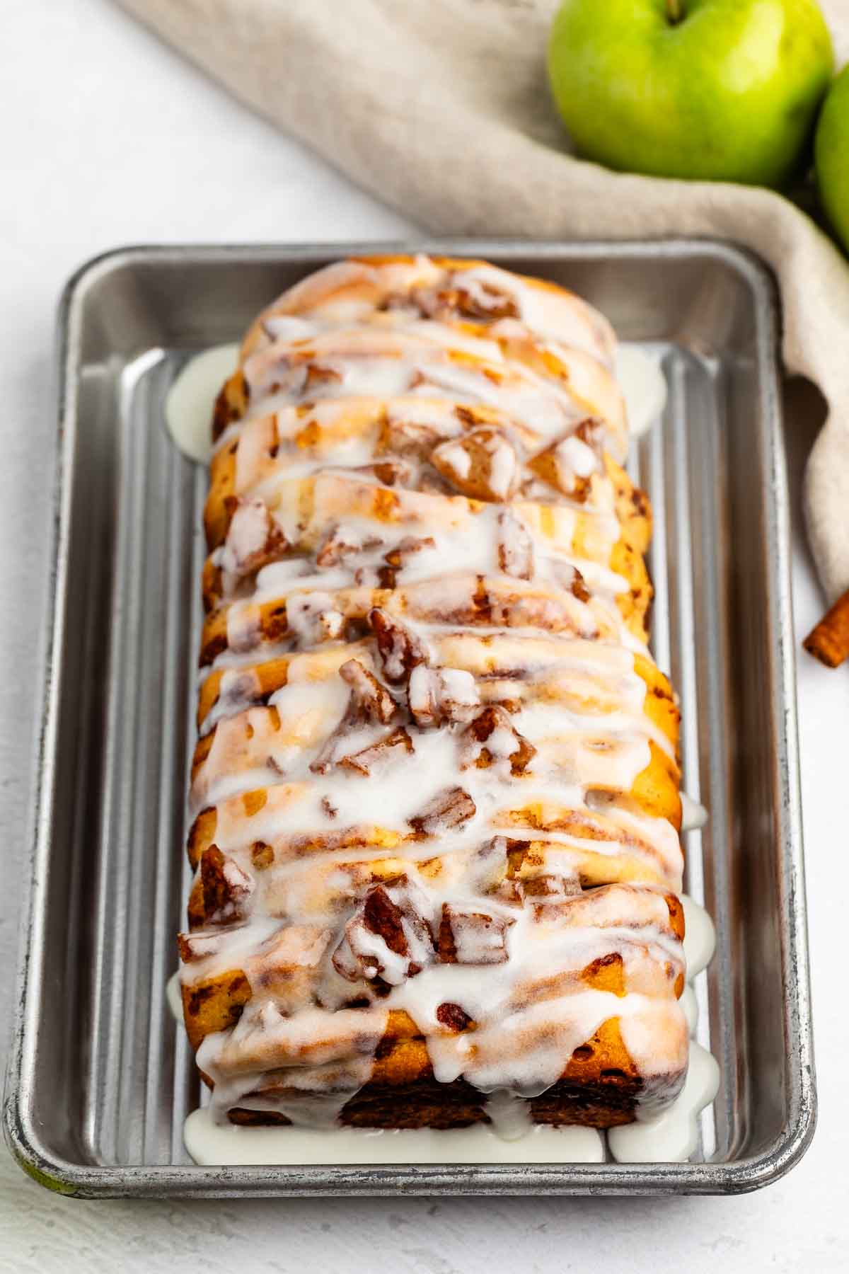 A loaf of apple cinnamon bread drizzled with white icing sits on a metal baking tray. In the background, a beige cloth and green apples are visible.
