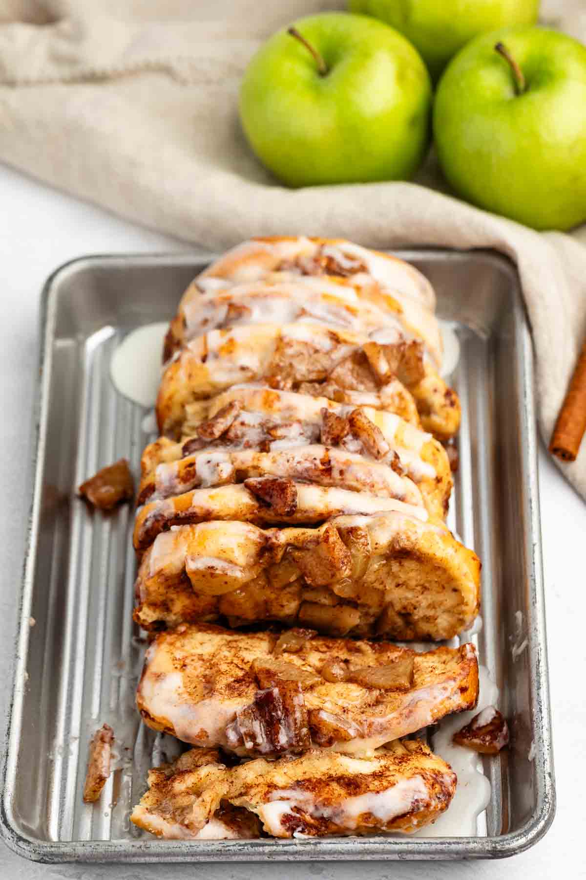 A sliced loaf of apple cinnamon bread drizzled with white icing sits on a metal tray. In the background, there are three green apples and a cinnamon stick on a beige cloth.