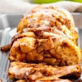 A close-up of sliced apple cinnamon bread topped with white icing, displayed on a metal tray with fresh green apples in the background.