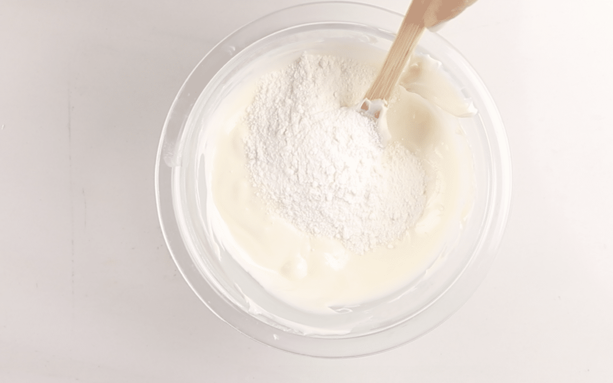 A hand holds a spatula, mixing white flour into a bowl of creamy batter on a white surface.