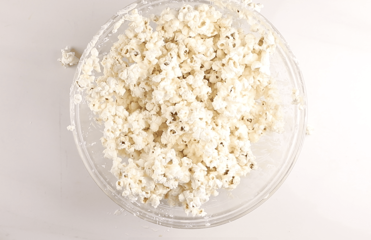 A clear glass bowl filled with freshly popped popcorn sits on a white surface, with a few pieces of popcorn scattered around the bowl.