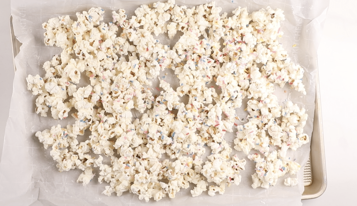 Popcorn covered in a white coating and colorful sprinkles spread out on a baking sheet lined with parchment paper.