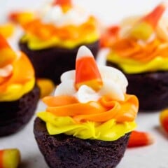 Several chocolate brownie bites topped with yellow and orange icing, a swirl of white frosting, and a piece of candy corn.