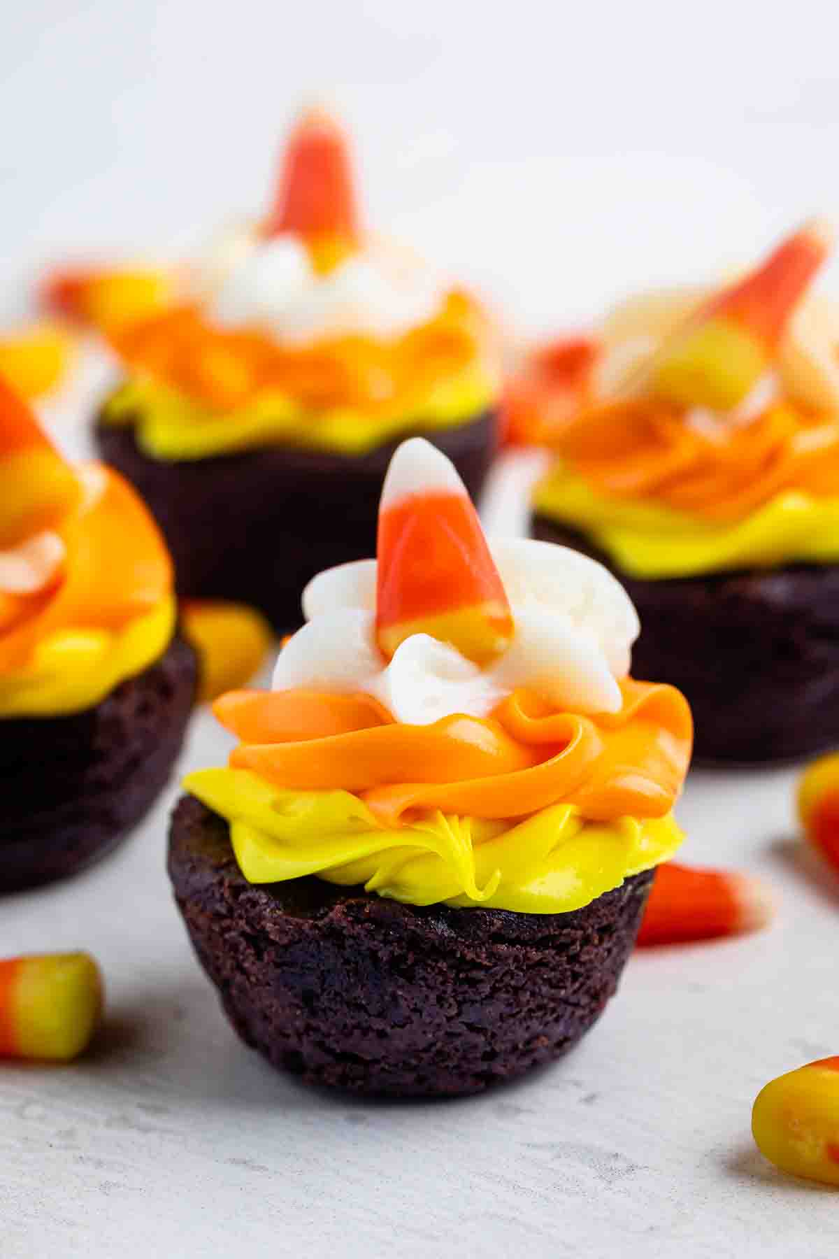 Several chocolate brownie bites topped with yellow and orange icing, a swirl of white frosting, and a piece of candy corn.