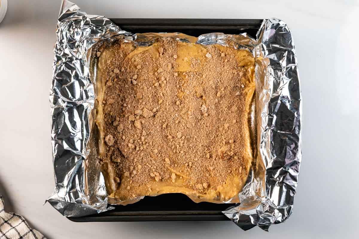 A square baking pan lined with crumpled aluminum foil, filled with an unbaked dessert topped with a crumbly brown mixture, sits on a white countertop.