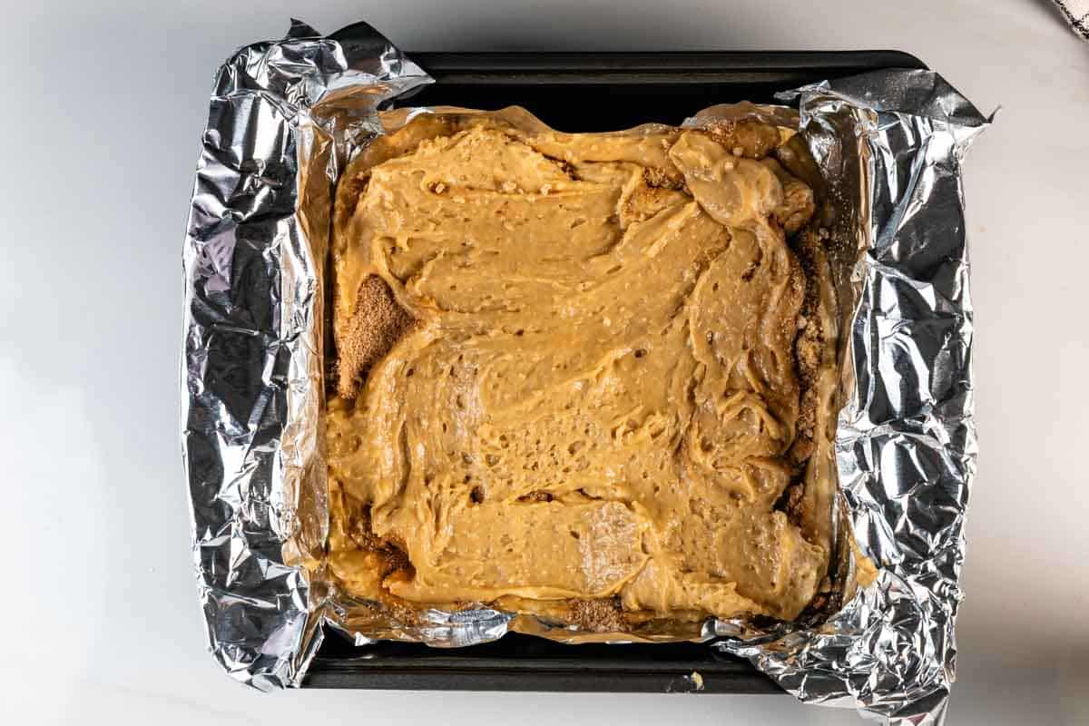 A square baking pan lined with crumpled aluminum foil holds a thick layer of creamy, light brown batter spread evenly on top. The pan sits on a white surface.