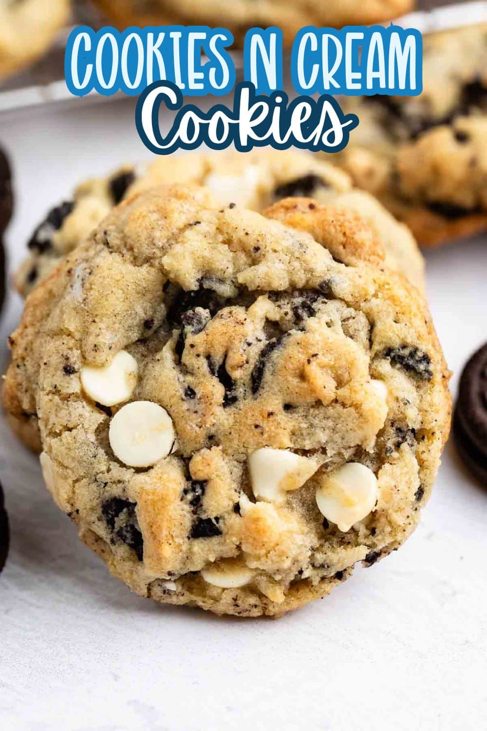 A close-up of a cookies and cream cookie with white chocolate chips and Oreo pieces, surrounded by more cookies and Oreos. Text at the top reads “Cookies N Cream Cookies.”.