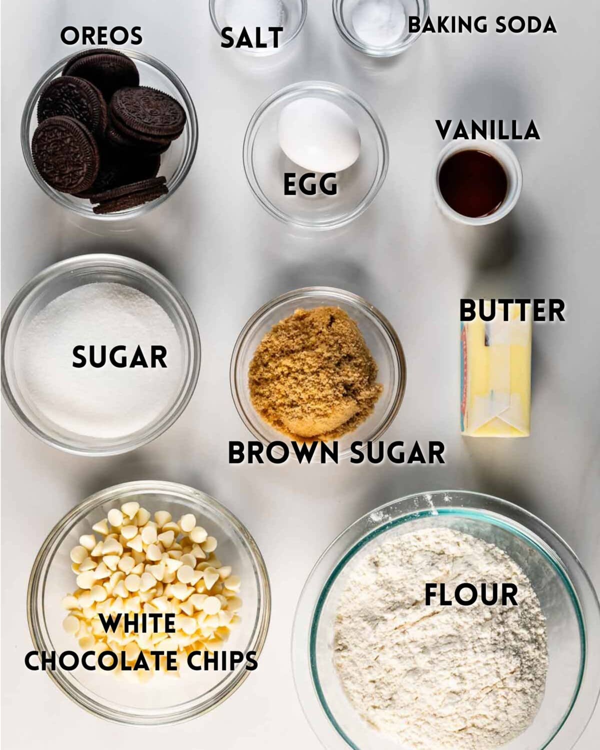 Overhead view of labeled baking ingredients in bowls: Oreos, salt, baking soda, egg, vanilla, sugar, brown sugar, butter, white chocolate chips, and flour, arranged neatly on a white surface.
