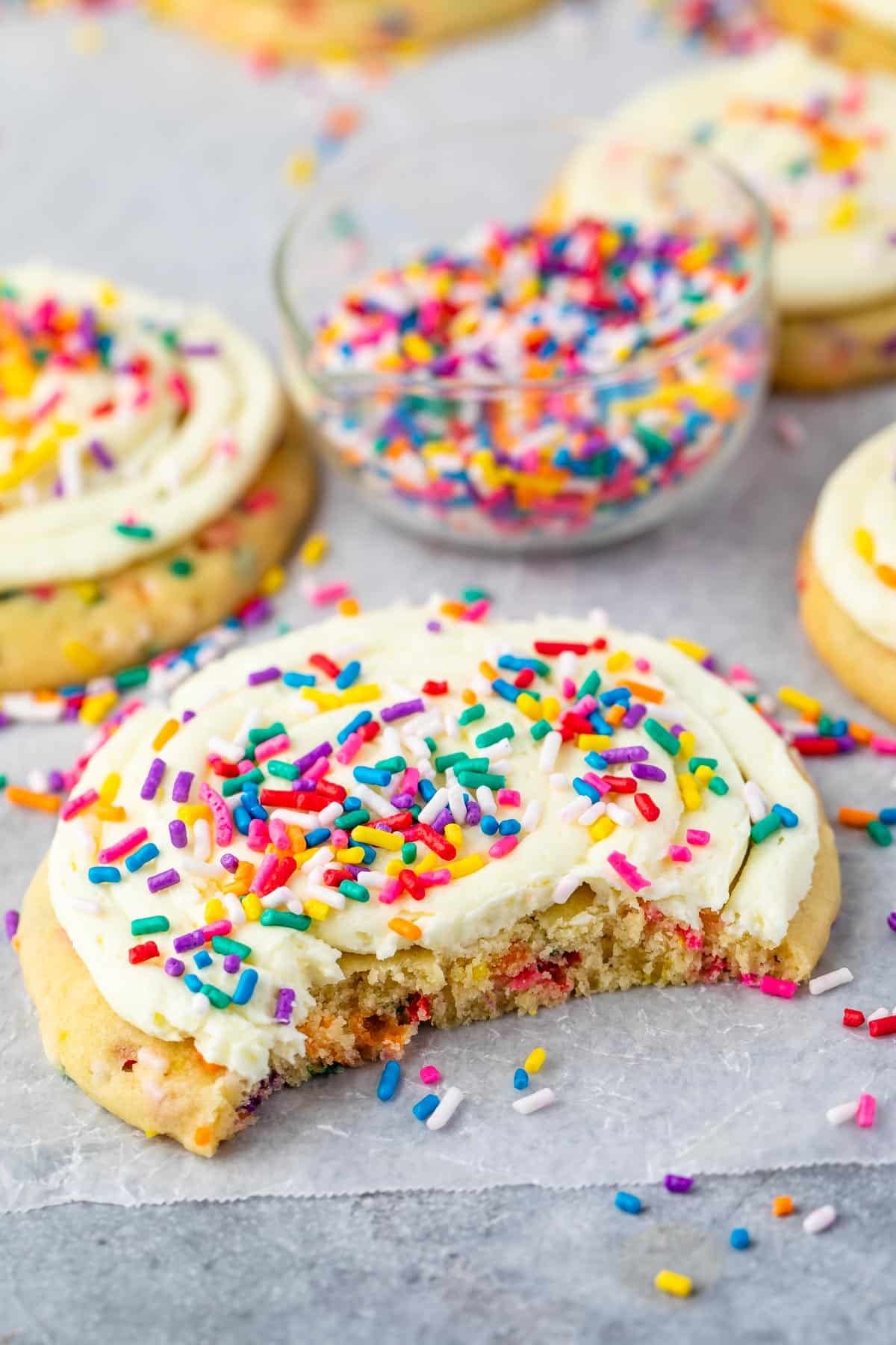 A frosted sugar cookie topped with colorful sprinkles sits on parchment paper, with a bite taken out. In the background, more cookies and a small glass bowl filled with rainbow sprinkles are visible.