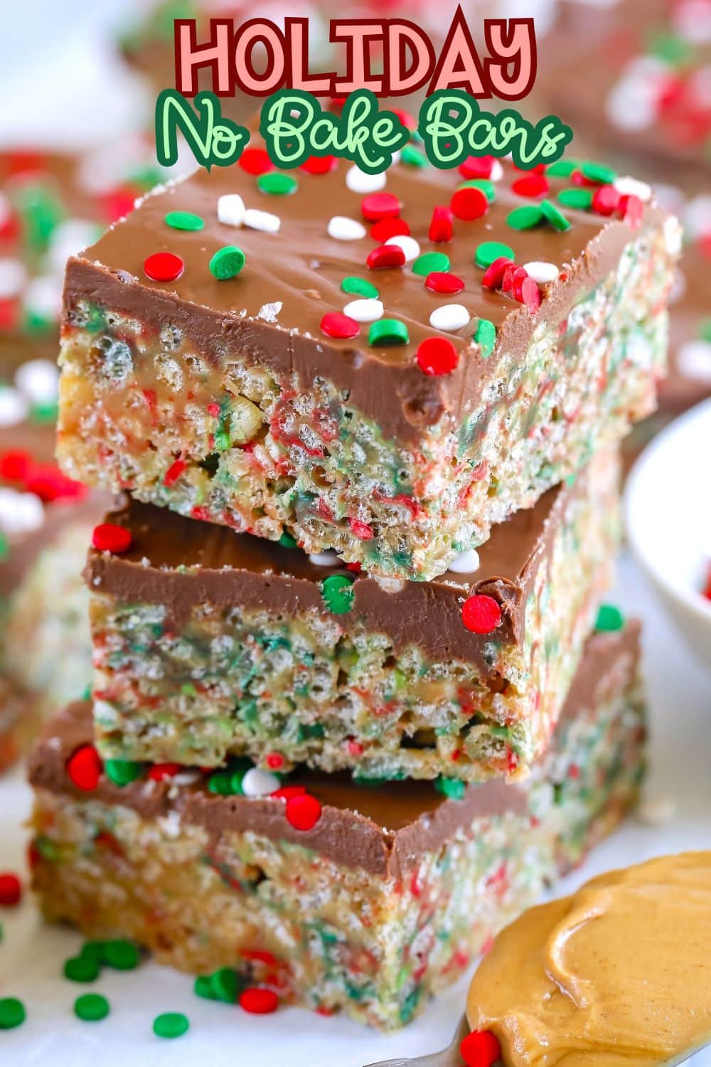 Three stacked no-bake dessert bars topped with chocolate and red, green, and white sprinkles. The bars have visible crispy rice cereal and are surrounded by more sprinkles and a spoonful of peanut butter. Text reads: Holiday No Bake Bars.