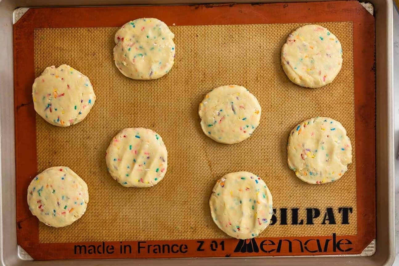 A baking sheet lined with parchment holds ten unbaked, round cookies with colorful sprinkles. Nearby are three boxes of Challenge Butter, two opened and one closed, on a white countertop.