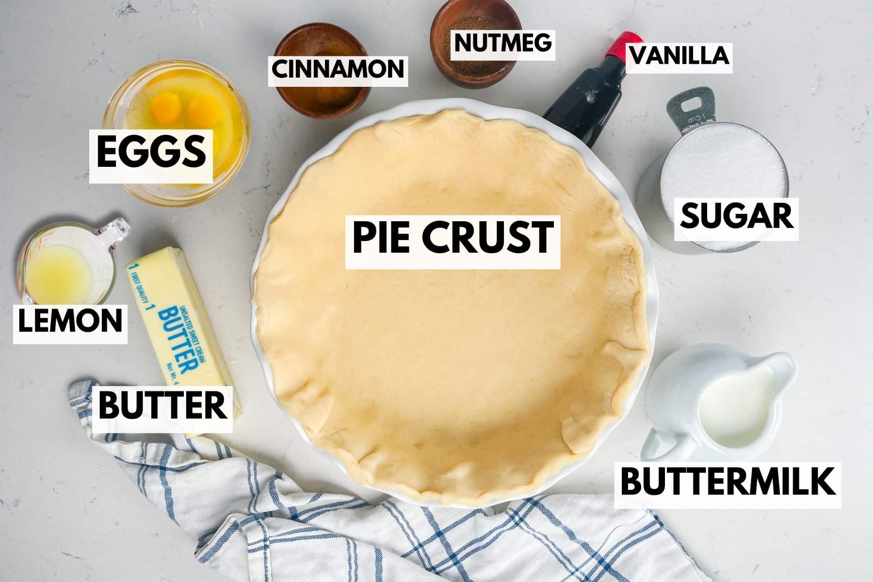 A pie crust in a dish is surrounded by labeled ingredients: eggs, lemon, butter, cinnamon, nutmeg, vanilla, sugar, and buttermilk, all arranged on a light countertop.