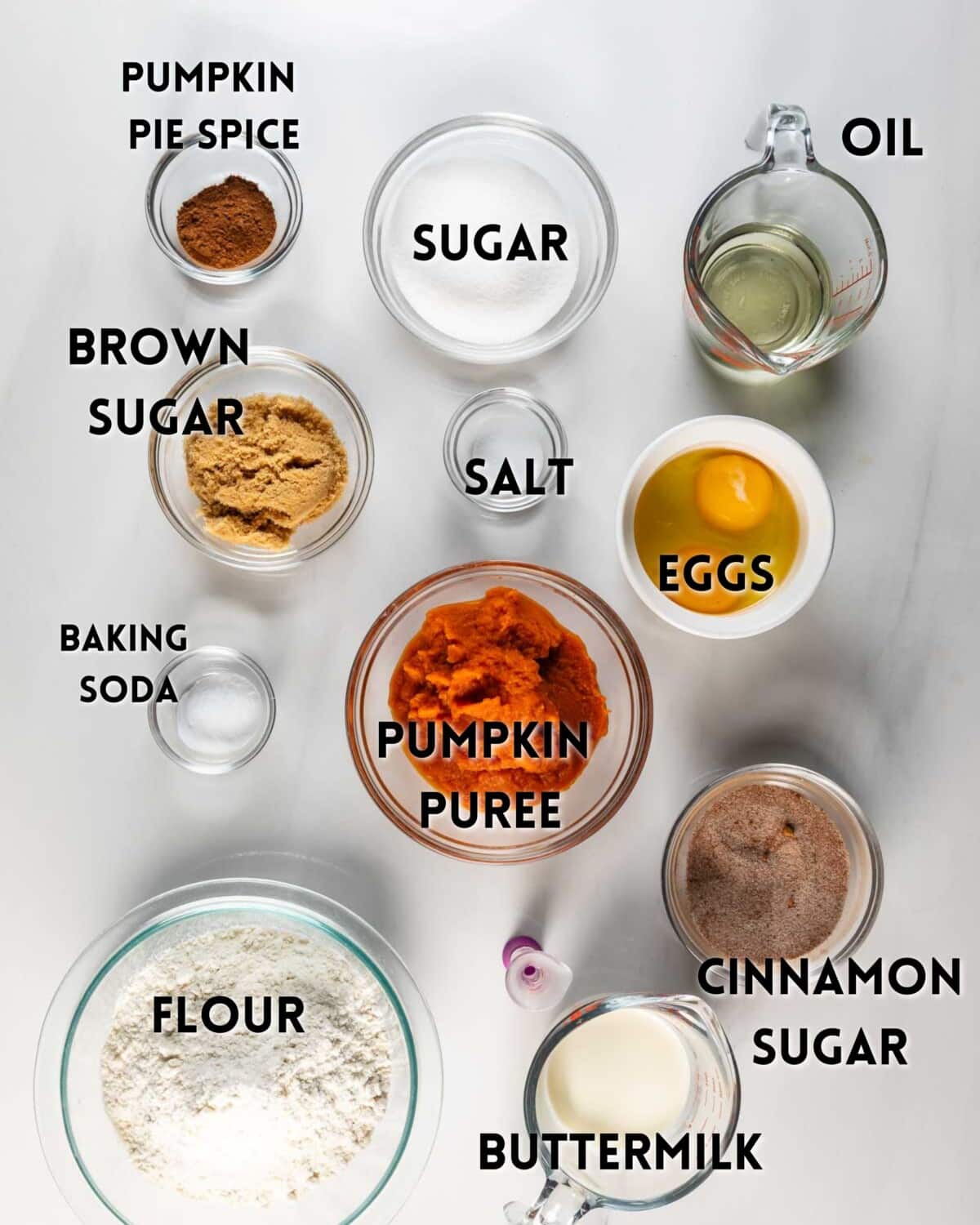 Various baking ingredients in bowls and measuring cups labeled: pumpkin pie spice, sugar, oil, eggs, salt, brown sugar, baking soda, pumpkin puree, cinnamon sugar, flour, and buttermilk, arranged on a white surface.