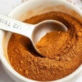 A metal tablespoon in a white bowl filled with brown ground spice, likely pumpkin pie spice or cinnamon, on a white surface with a small orange decoration and cinnamon stick nearby.