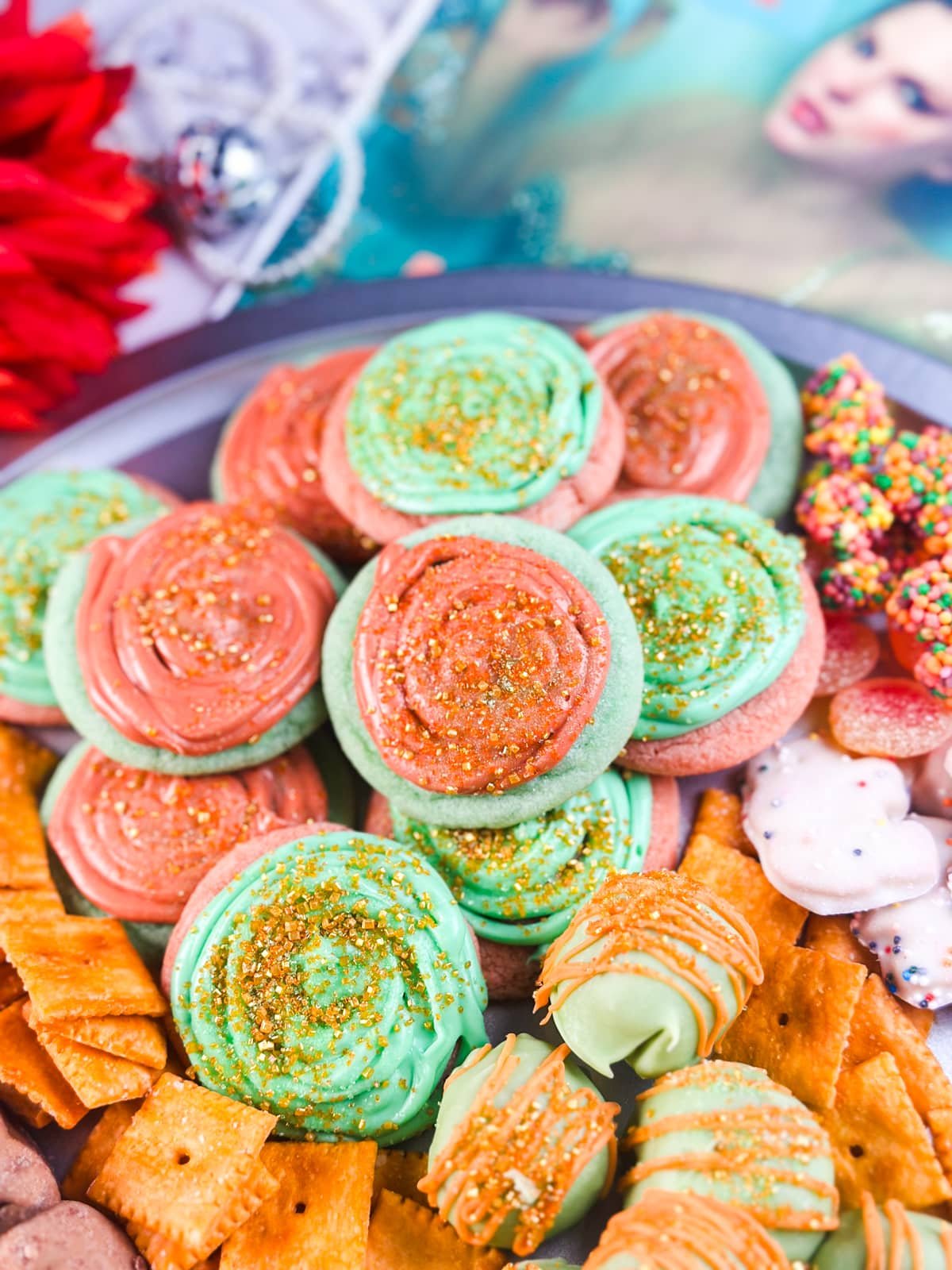 A close-up of a platter filled with frosted swirl cookies in green and red, sprinkled with gold, surrounded by crackers, chocolate-covered snacks, and other treats. A blurred magazine and decor are in the background.