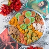 A round tray filled with colorful cookies, crackers, animal-shaped snacks, and candies sits on a white surface. Red flowers, sparkling beads, disco balls, and two Bollywood magazine covers frame the tray.