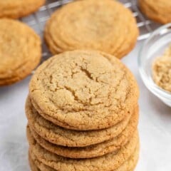 A stack of six golden brown cookies sits in the foreground, with more cookies and a cooling rack in the background. A small glass bowl with brown sugar is partially visible to the right.
