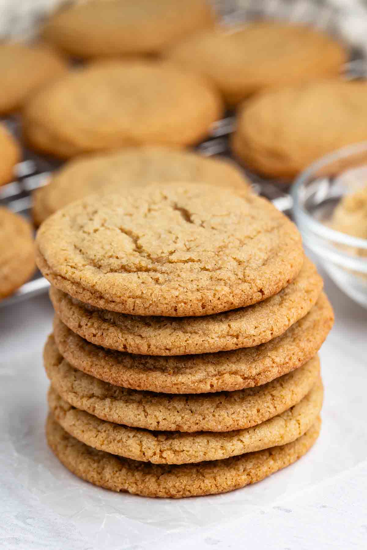 A stack of six golden brown, round cookies sits on a white surface, with more cookies and a glass bowl of brown sugar blurred in the background.