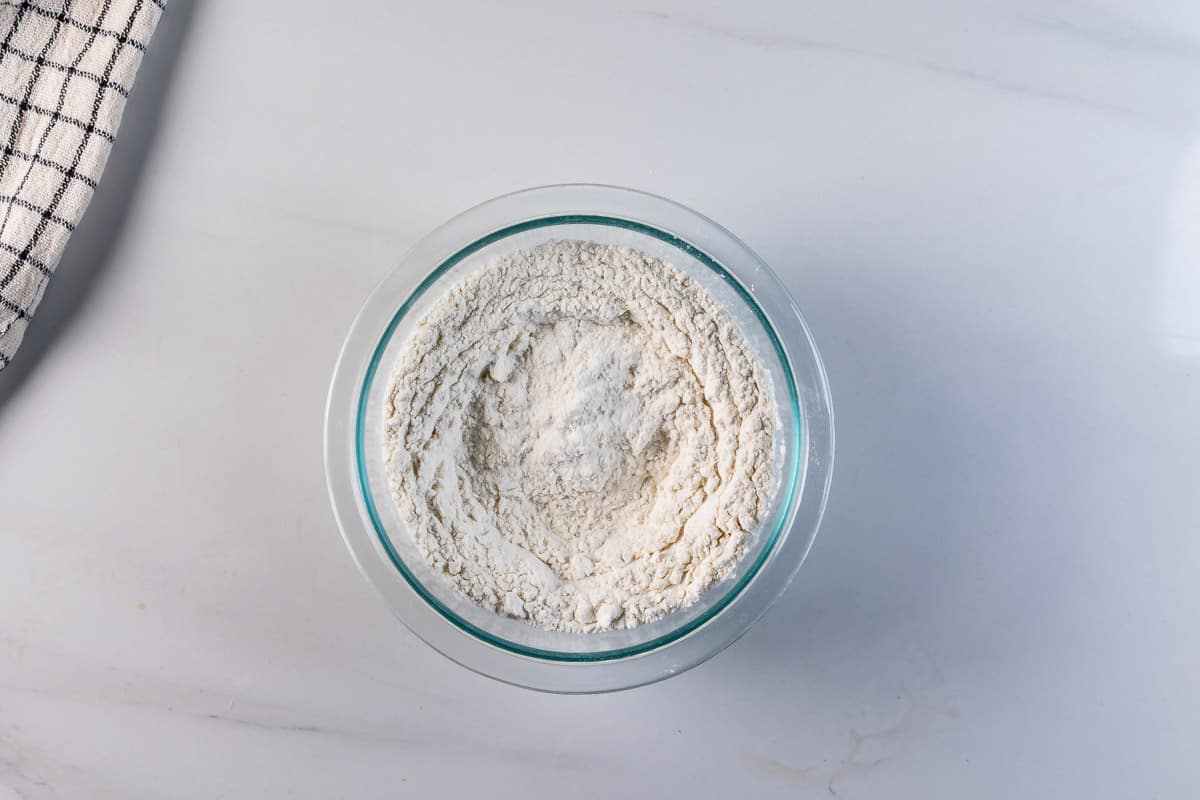 A glass bowl filled with flour sits on a white marble surface; a checkered cloth is partially visible in the top left corner.