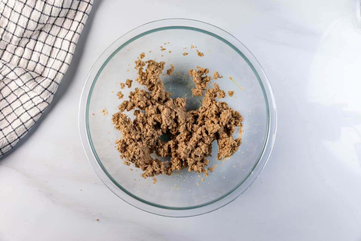 A glass bowl containing a mixture of brown cookie dough sits on a white marble surface next to a black-and-white checkered kitchen towel.