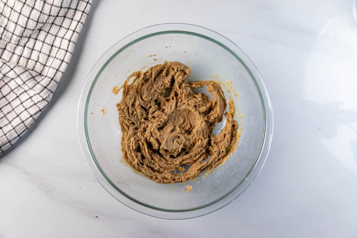 A glass mixing bowl with brown cookie dough sits on a white countertop beside a black-and-white checkered kitchen towel.