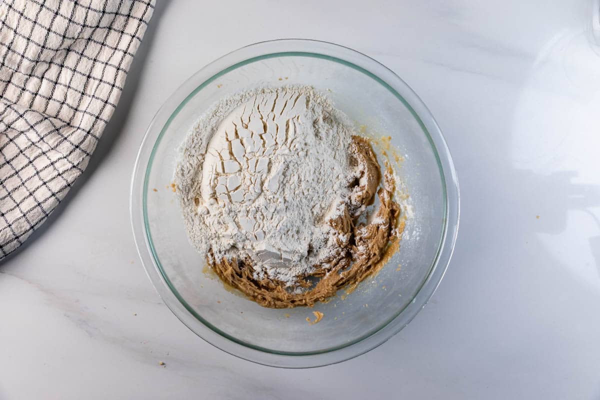 A glass mixing bowl containing partially mixed cookie dough with a mound of flour on top sits on a white countertop next to a black and white checkered kitchen towel.