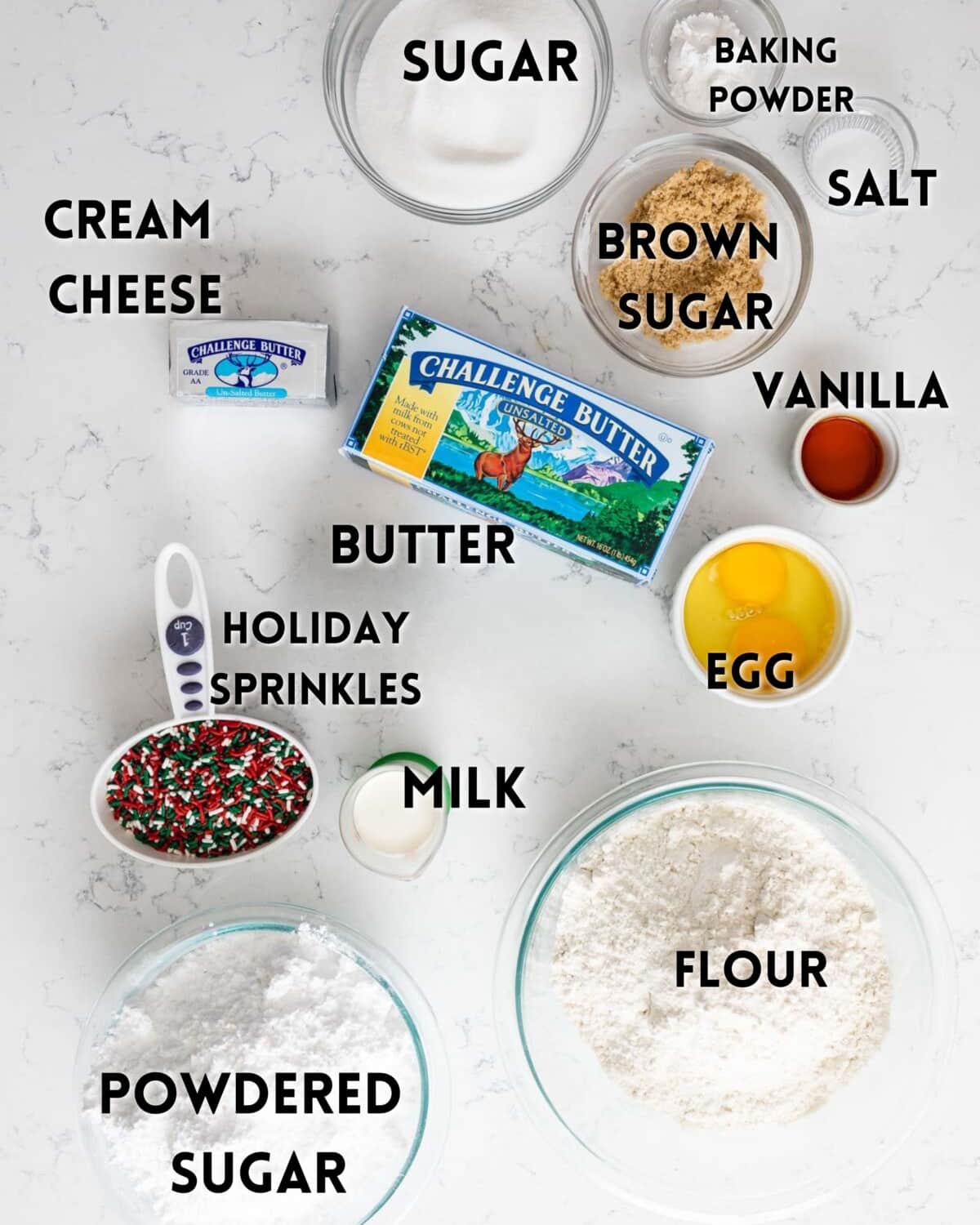 Labeled baking ingredients on a white surface: sugar, baking powder, brown sugar, salt, vanilla, cream cheese, butter, egg, holiday sprinkles, milk, flour, and powdered sugar.