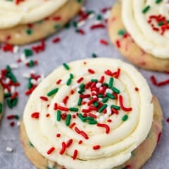 A close-up of a frosted sugar cookie topped with swirled vanilla icing and red, green, and white sprinkles, set on a parchment paper background with other cookies nearby.