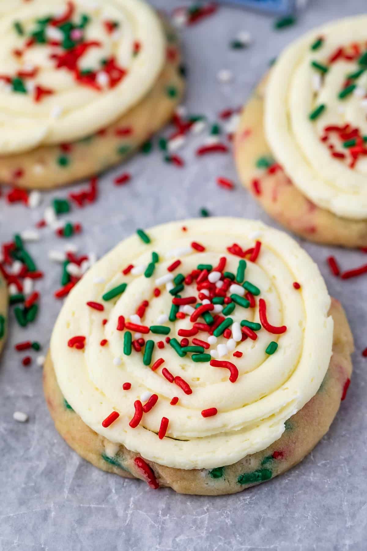 A close-up of a frosted sugar cookie topped with swirled vanilla icing and red, green, and white sprinkles, set on a parchment paper background with other cookies nearby.