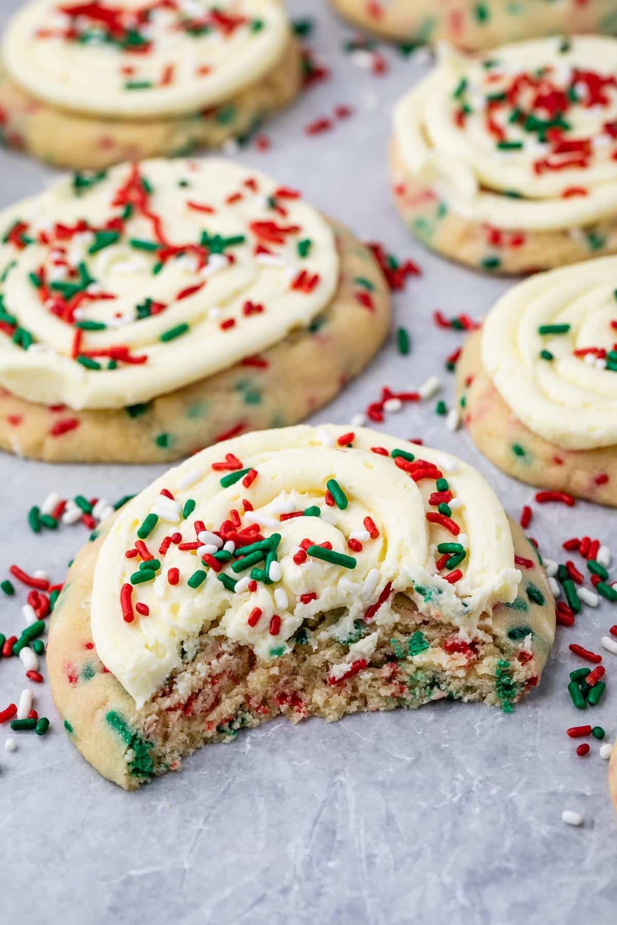 A frosted sugar cookie with red and green sprinkles sits on a surface, with a bite taken out of it. Other similar cookies and scattered sprinkles are visible in the background.
