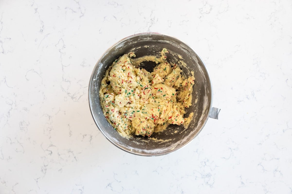 A metal mixing bowl filled with cookie dough mixed with colorful sprinkles sits on a white, marbled countertop.