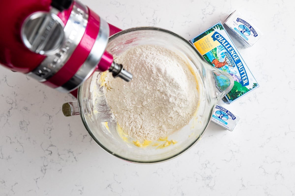 A stand mixer with a bowl containing flour and butter sits on a white countertop, next to two sticks of Challenge brand butter.