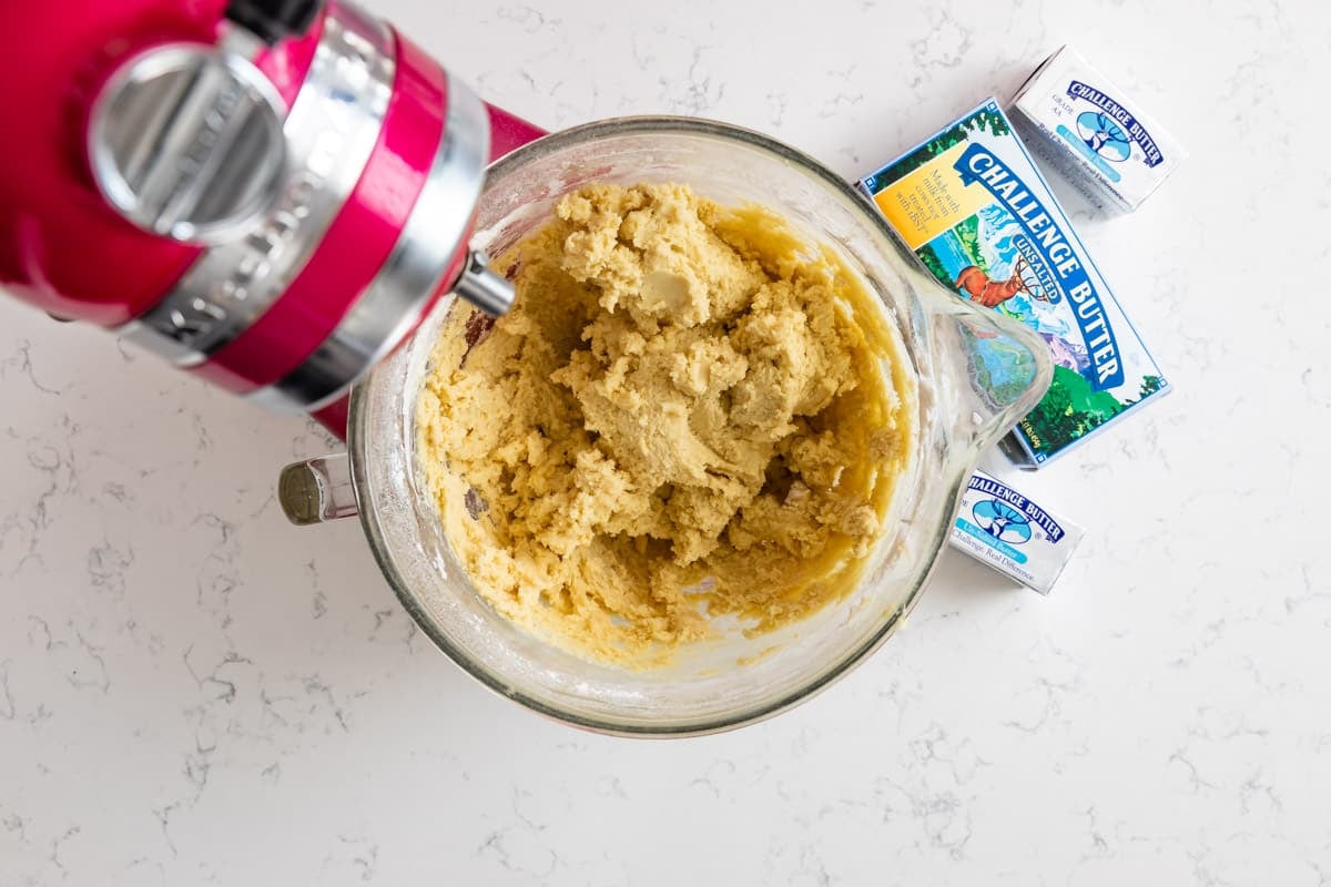A stand mixer with cookie dough in a glass bowl sits on a white countertop next to two packs of Challenge Butter.