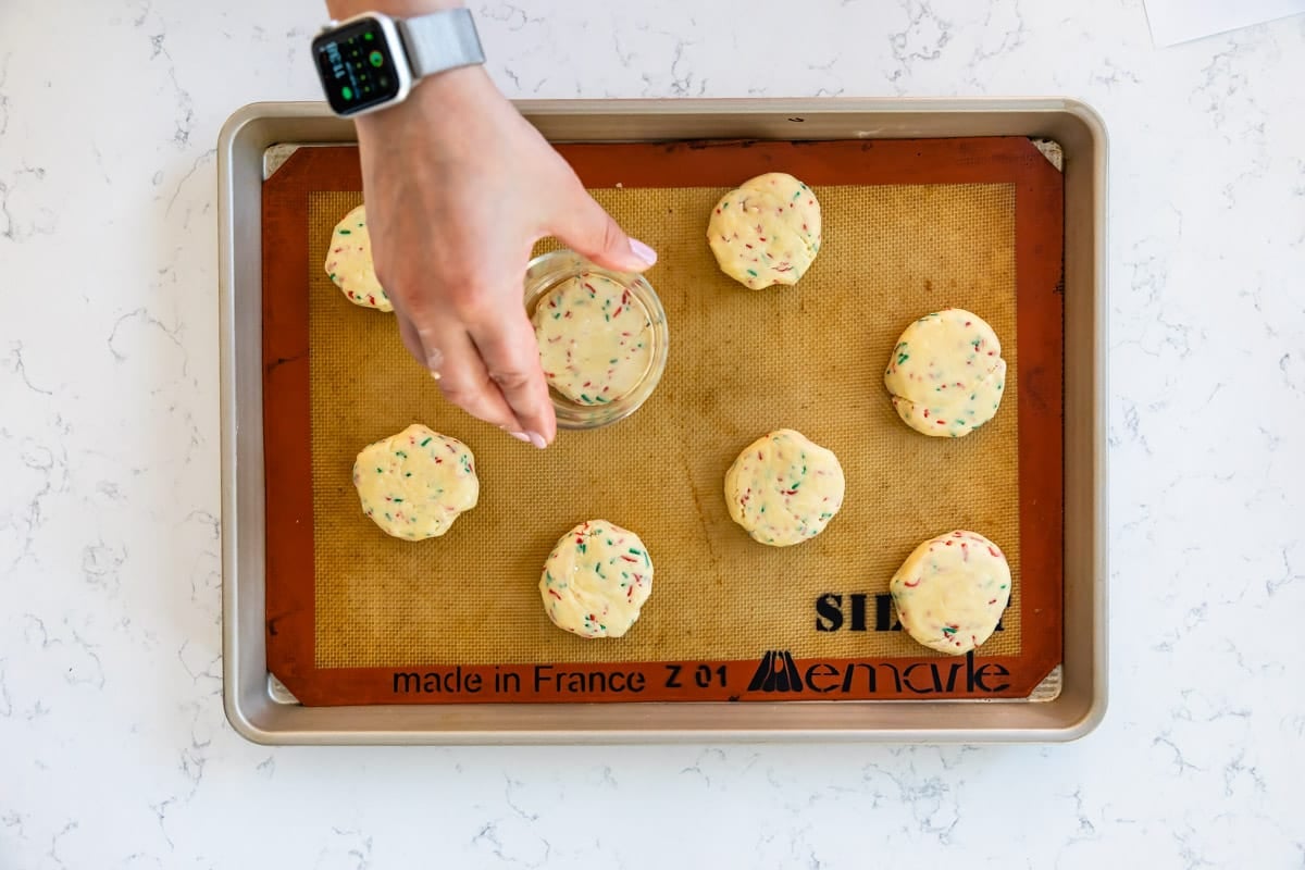 A hand pressing down cookie dough with a glass on a baking tray lined with a silicone mat. The dough has colorful sprinkles and is arranged in rows, ready to be baked. The person is wearing a smartwatch.
