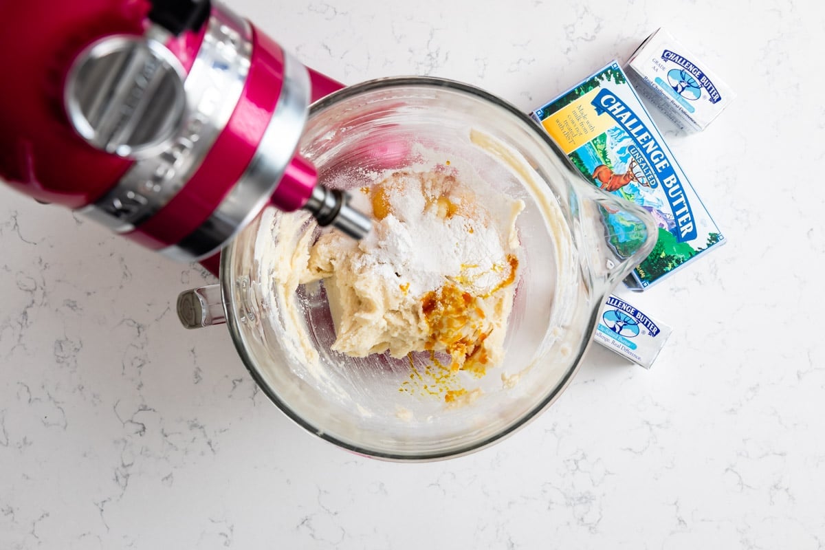 A stand mixer with a glass bowl is blending dough with visible flour and orange zest. Next to the mixer are two packs of Challenge unsalted butter on a white marble countertop.