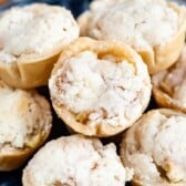 A close-up view of several mini apple pies with golden, crumbly tops and flaky crusts, arranged on a dark plate.