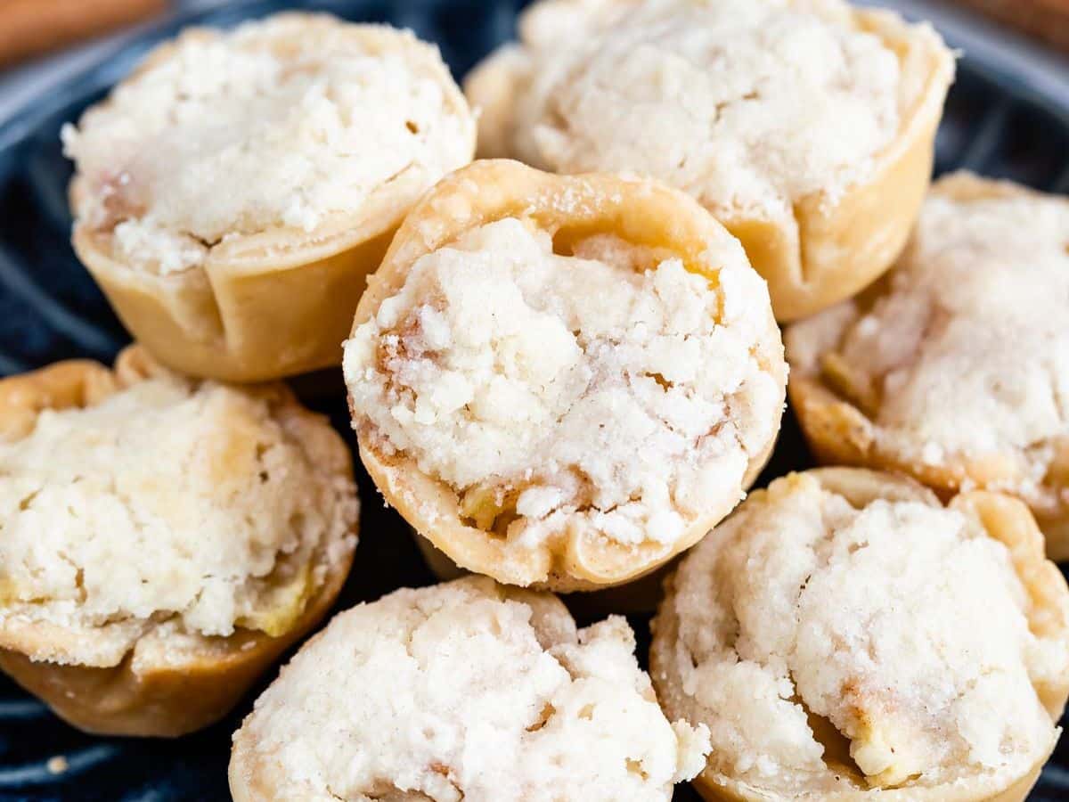 A close-up view of several mini apple pies with golden, crumbly tops and flaky crusts, arranged on a dark plate.