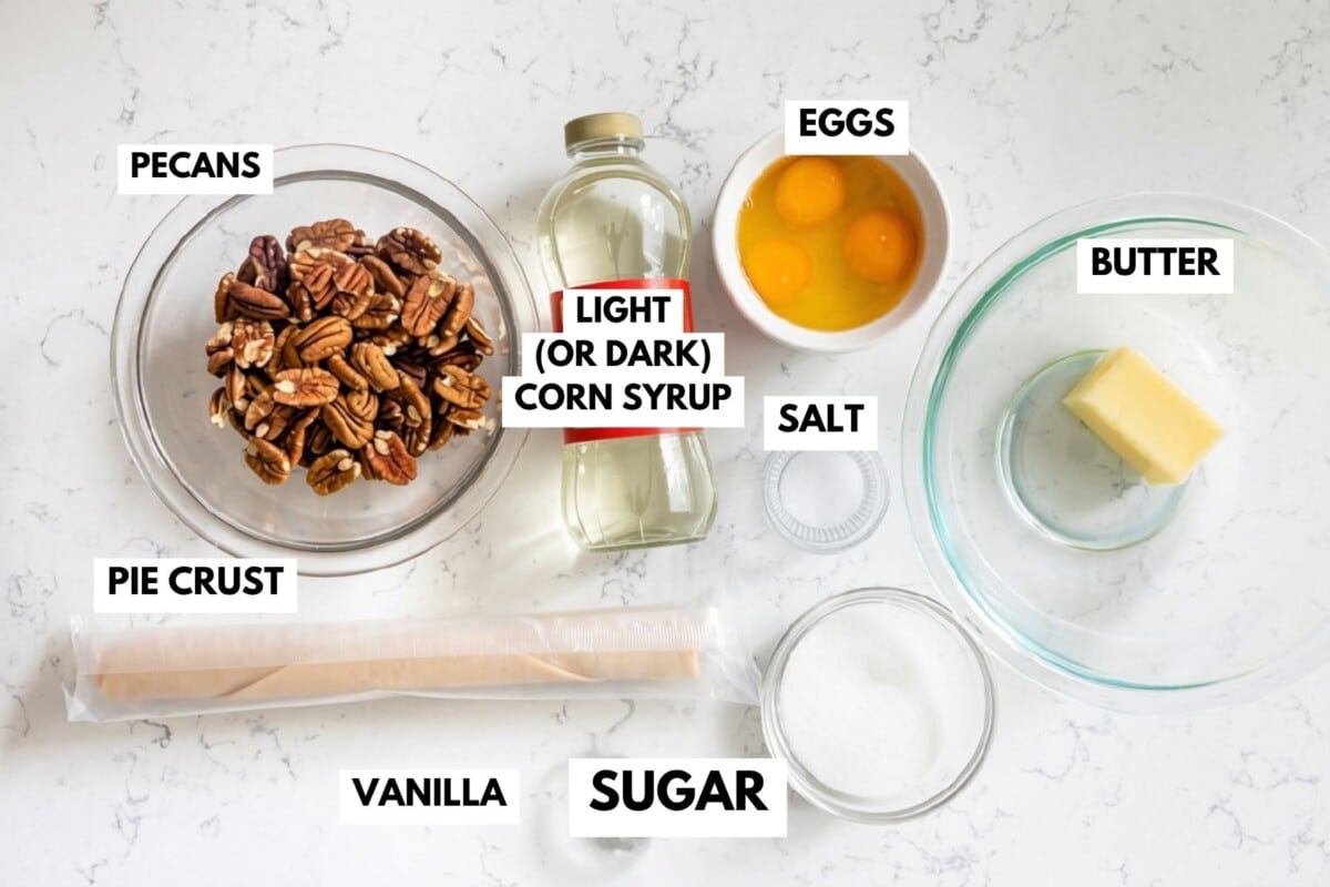 Labeled baking ingredients on a countertop for mini pecan pies: pecans, pie crust, vanilla, light (or dark) corn syrup, eggs, salt, butter, and sugar, each in separate bowls or containers.