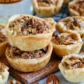 Mini pecan pies with flaky crusts are stacked on a wooden board, surrounded by more mini pecan pies and scattered pecan pieces. The pies have a golden-brown color and a crunchy pecan topping.