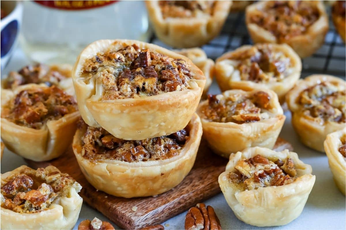 Mini pecan pies with flaky crusts are stacked on a wooden board, surrounded by more mini pecan pies and scattered pecan pieces. The pies have a golden-brown color and a crunchy pecan topping.