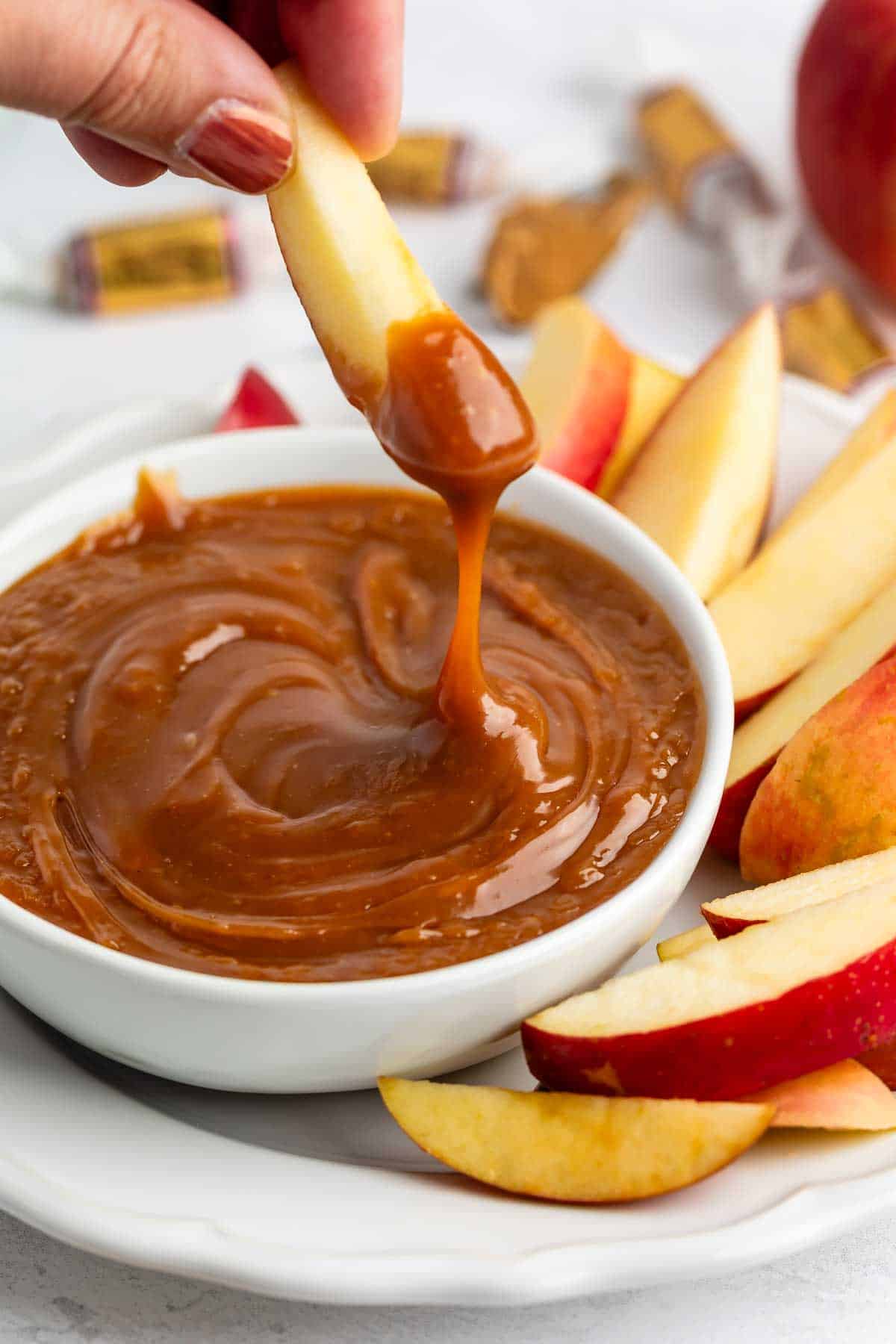 A hand dips a fresh apple slice into a bowl of creamy caramel sauce, surrounded by more apple slices on a white plate. Wrapped caramel candies are visible in the background.