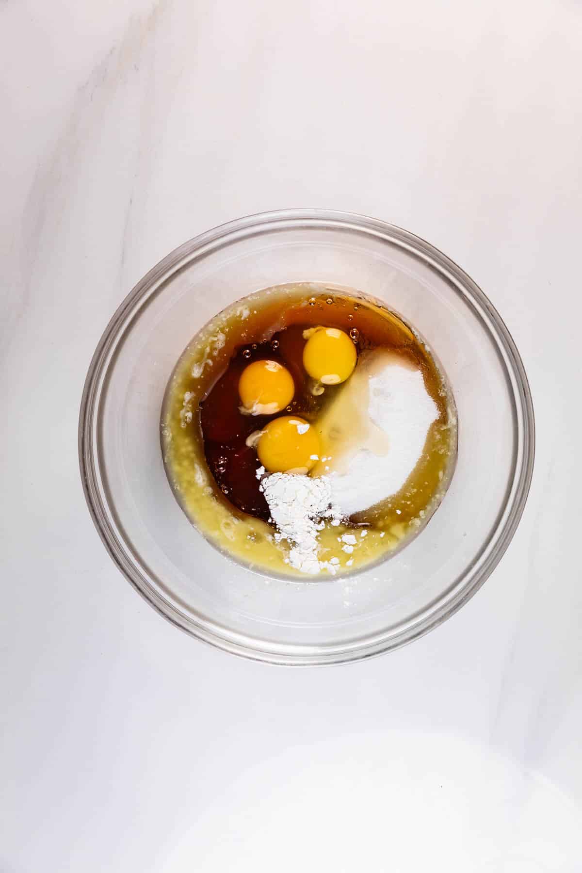 A glass mixing bowl containing three raw eggs, sugar, flour, vanilla extract, and other baking ingredients on a white marble surface.