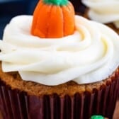 A pumpkin cupcake with white frosting in a swirl pattern, topped with a small candy pumpkin, in a brown paper liner.