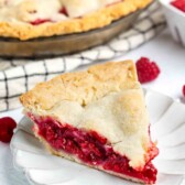 A slice of raspberry pie with a golden, flaky crust sits on a white plate. The filling is bright red and juicy, and the rest of the pie is visible in the background alongside fresh raspberries.