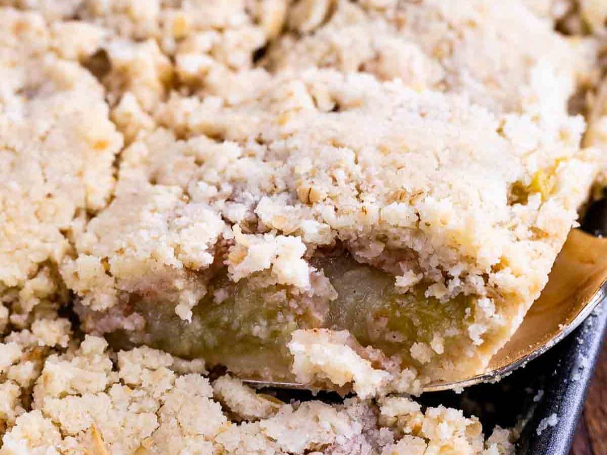 A close-up of a crumb-topped fruit dessert, reminiscent of an Apple Slab Pie, with a golden, crumbly topping and juicy fruit filling visible as a piece is lifted from the baking pan.