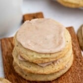 A stack of three frosted cookies with light brown icing sits on a small wooden board. More cookies and a white mug are visible in the background.