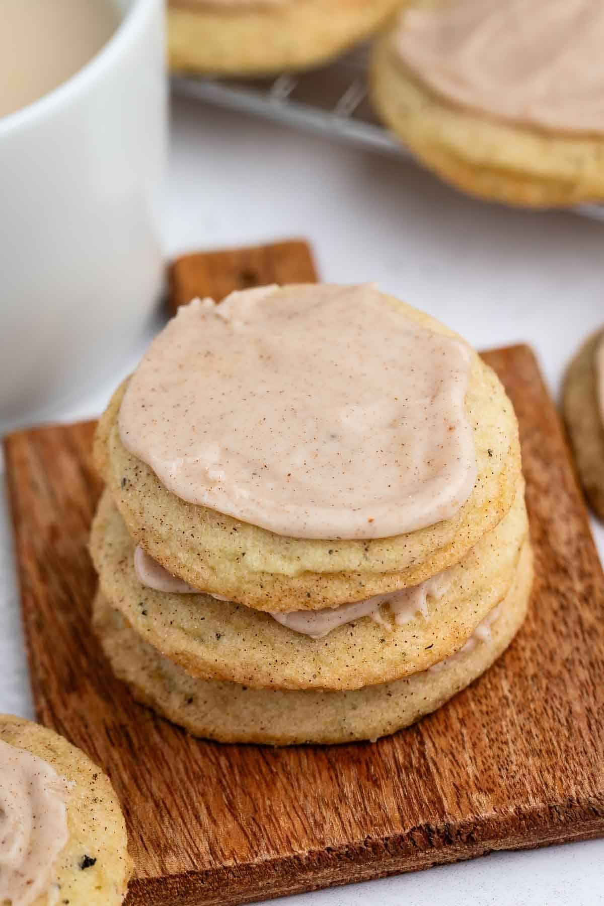 A stack of three frosted cookies with light brown icing sits on a small wooden board. More cookies and a white mug are visible in the background.