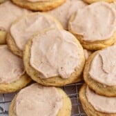 A close-up of soft, round cookies topped with a light brown frosting, arranged on a cooling rack with more cookies in the background.