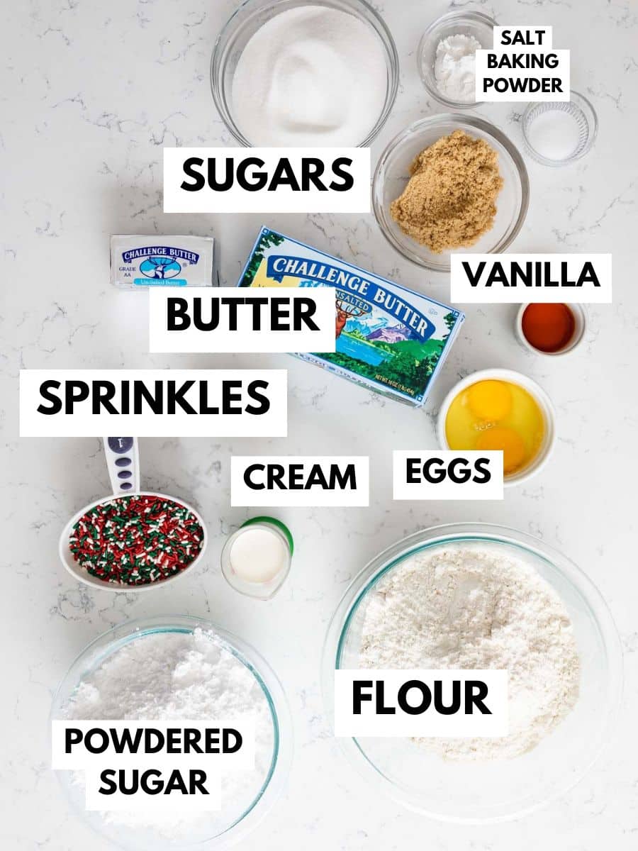 A top-down view of baking ingredients for Christmas cake batter cookies labeled on a white surface: sugars, vanilla, salt, baking powder, butter, sprinkles, cream, eggs, powdered sugar, and flour in bowls and containers.